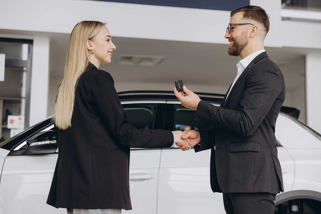 A young couple sits at a desk with paperwork and car keys in front of them, discussing terms with a financial advisor.