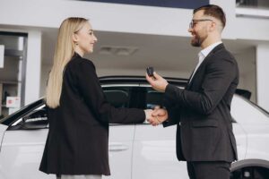A young couple sits at a desk with paperwork and car keys in front of them, discussing terms with a financial advisor.
