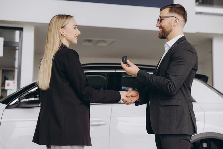 A young couple sits at a desk with paperwork and car keys in front of them, discussing terms with a financial advisor.